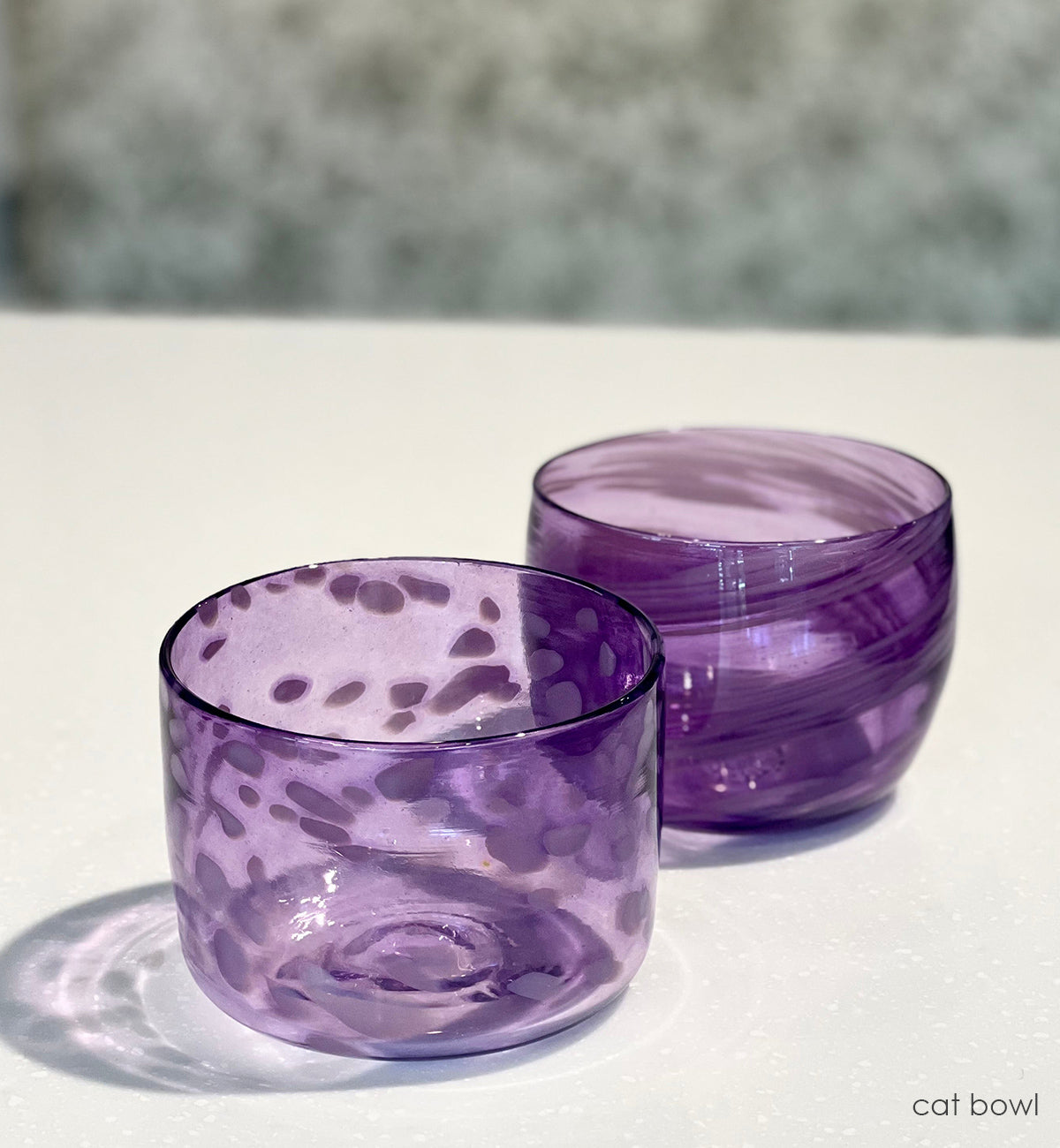 Two purple glass cat bowls on a white surface with a blurred background