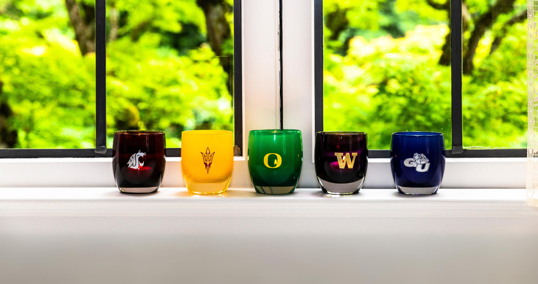 etched hand-blown glass candle holders with a variety of collegiate logos sitting on a window seal.