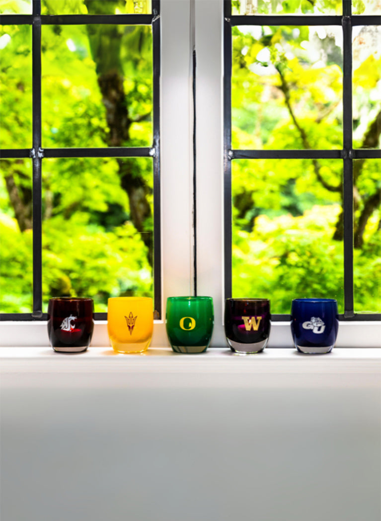 etched hand-blown glass candle holders with a variety of collegiate logos sitting on a window seal.