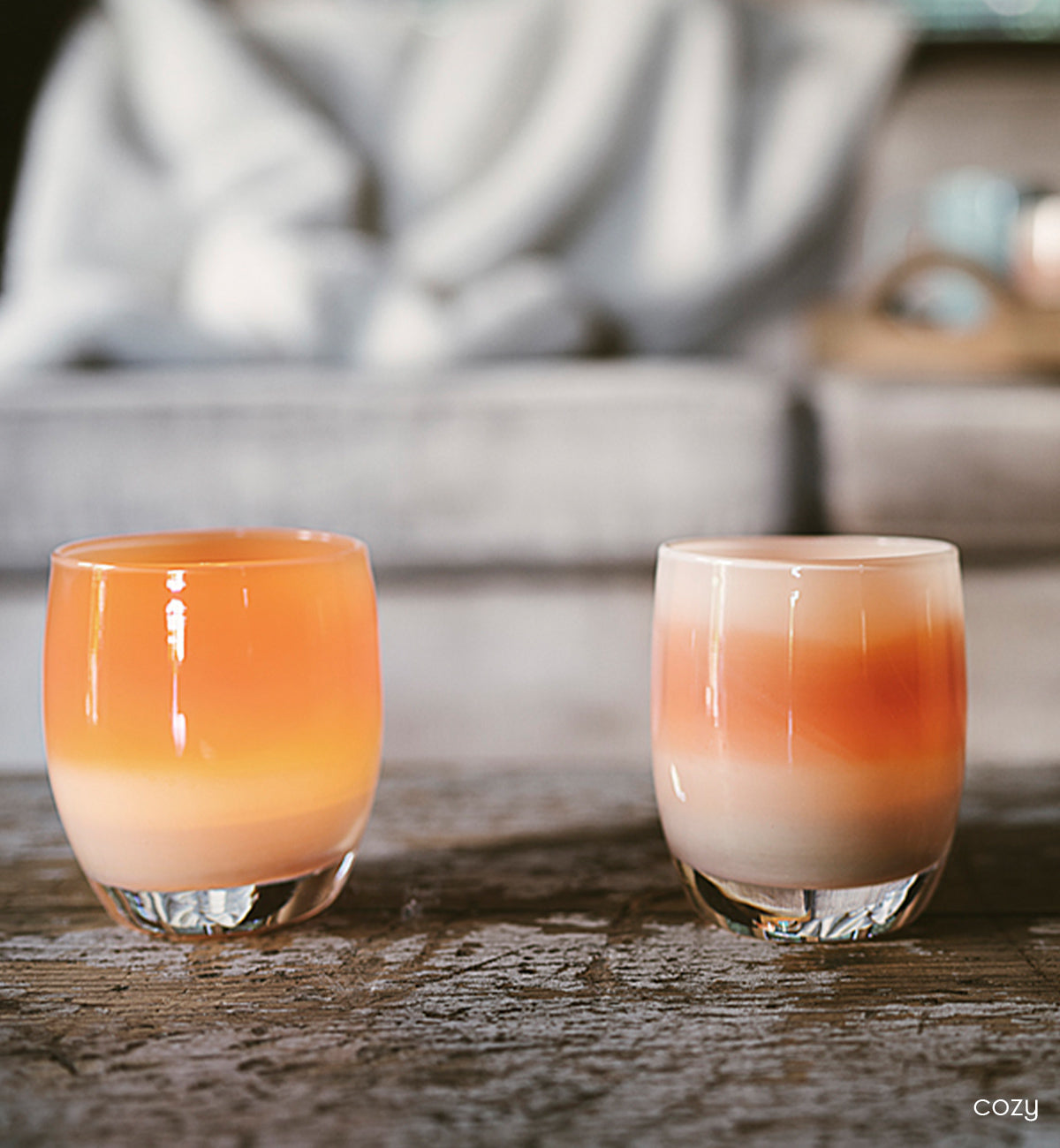 cozy, two apricot orange stripey hand-blown glass votive candle holders on a wood coffee table with a gray cozy couch in background.