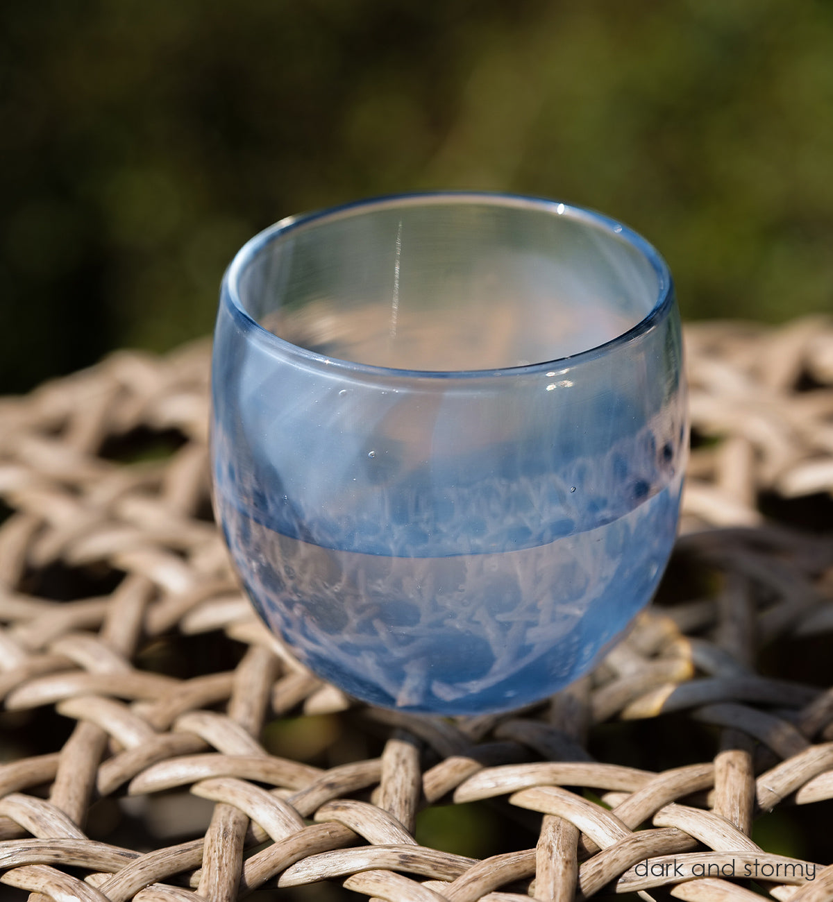 dark and stormy deep blue and white swirled  hand-blown drinking glass sitting on a wicker table outside.
