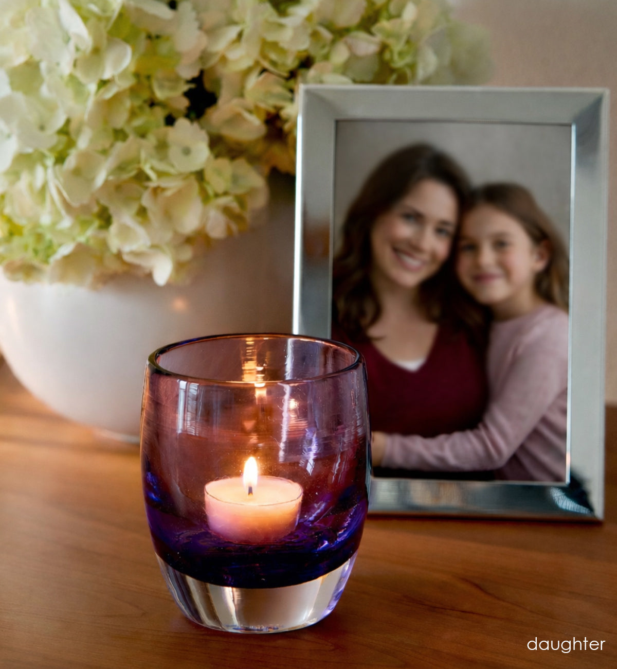 'daughter' lit purple hand-blown glass candle holder on wood dresser. Photo of  mother and daughter in silver photo frame and flowers in white vase in background.
