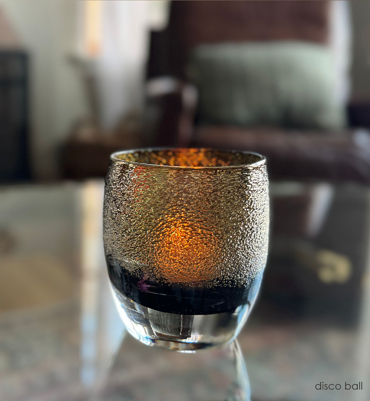 disco ball, black and gold shimmer hand-blown glass votive candle holder on a clear glass coffee table with chair in background.