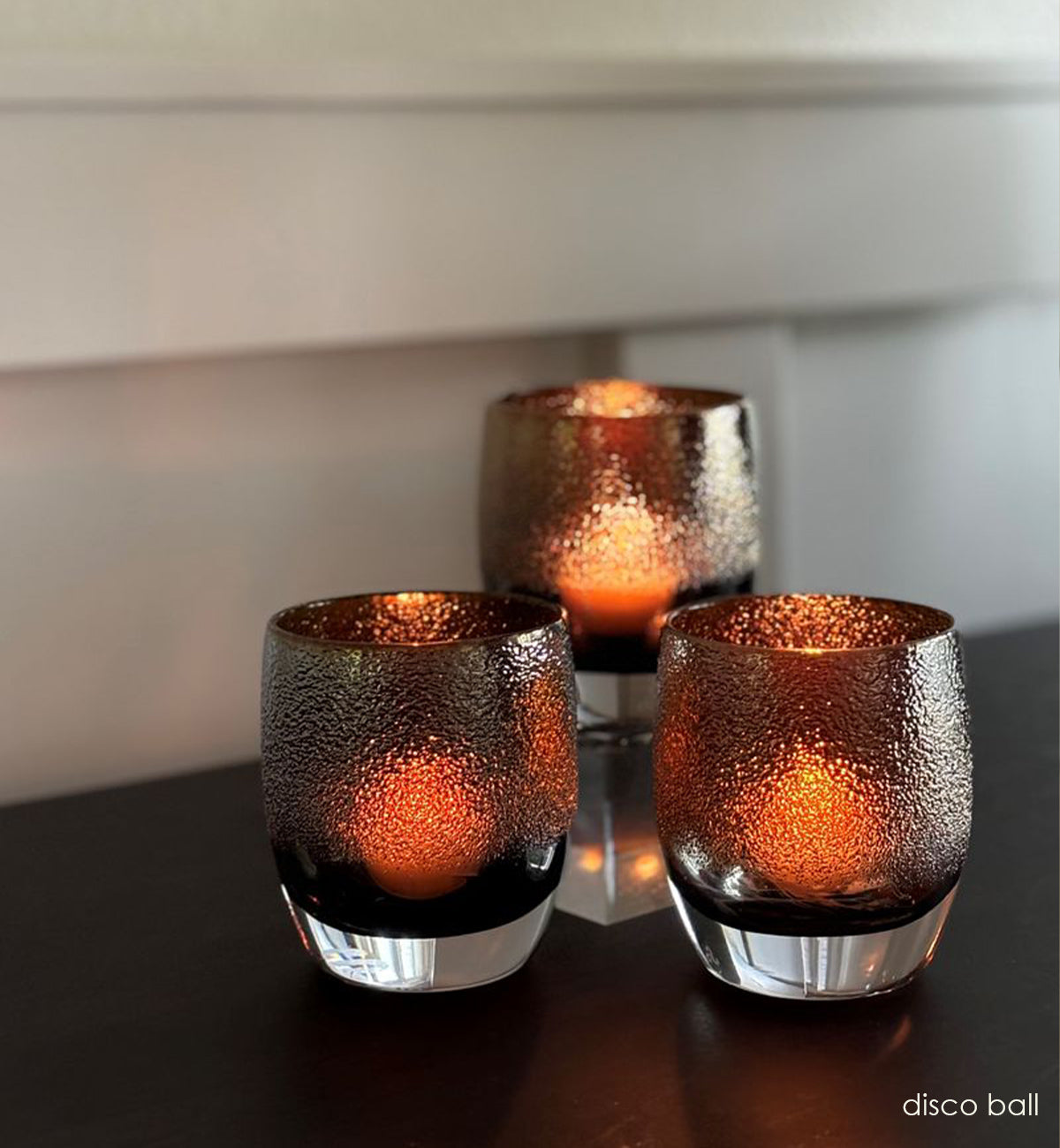 three disco ball, translucent black with gold shimmer hand-blown glass votive candle holders with lit tealights glowing through inside on a dark wood table against a white wall, with one votive on a square baby stand.