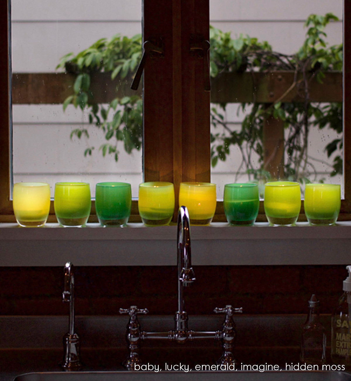 emerald, green hand-blown glass votive candle holder iwth baby, lucky, imagine and hidden moss on a kitchen sink windowsill.
