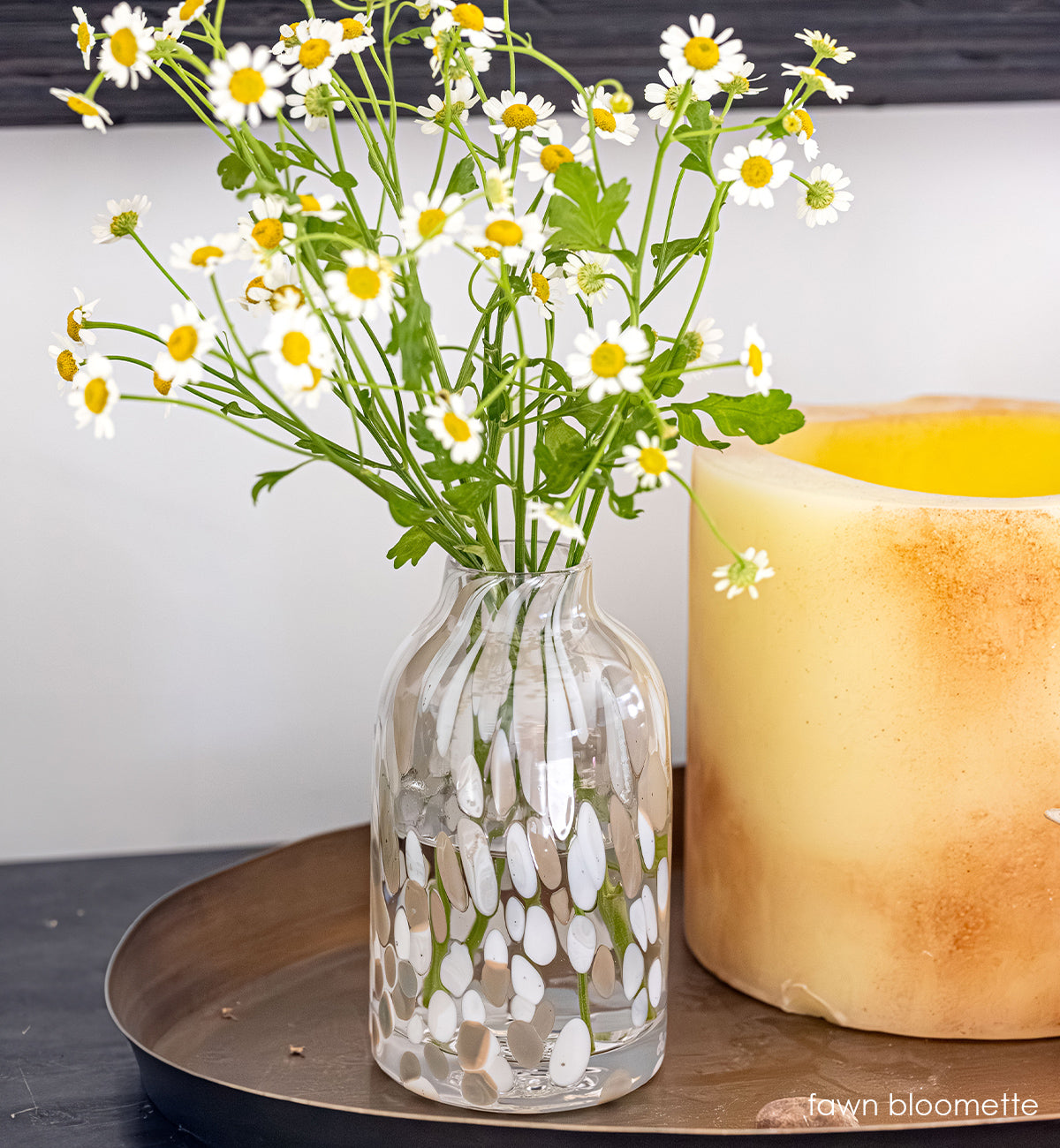 clear bloomette  vase with white and tan specs holding a bouquet of white and yellow flowers on a wooden surface with a candle in the background.