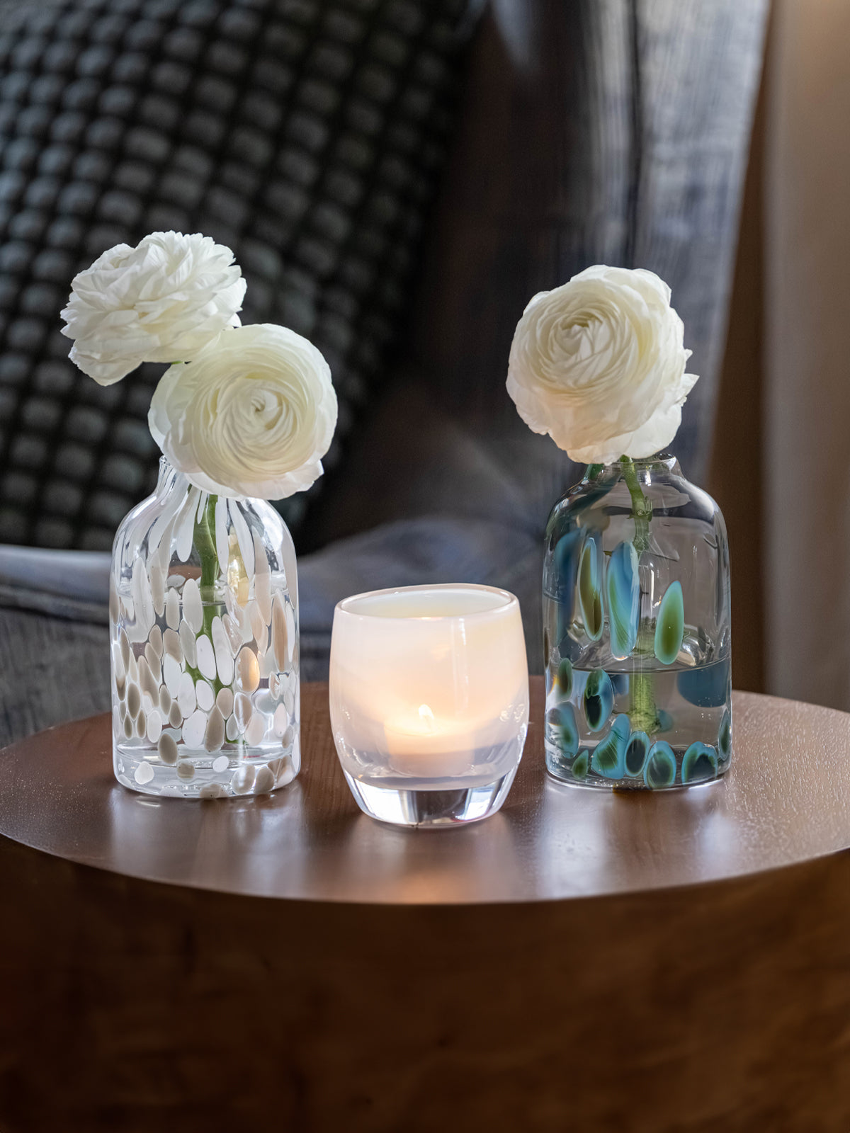 Two glass vases with white flowers and a lit candle on a wooden surface.