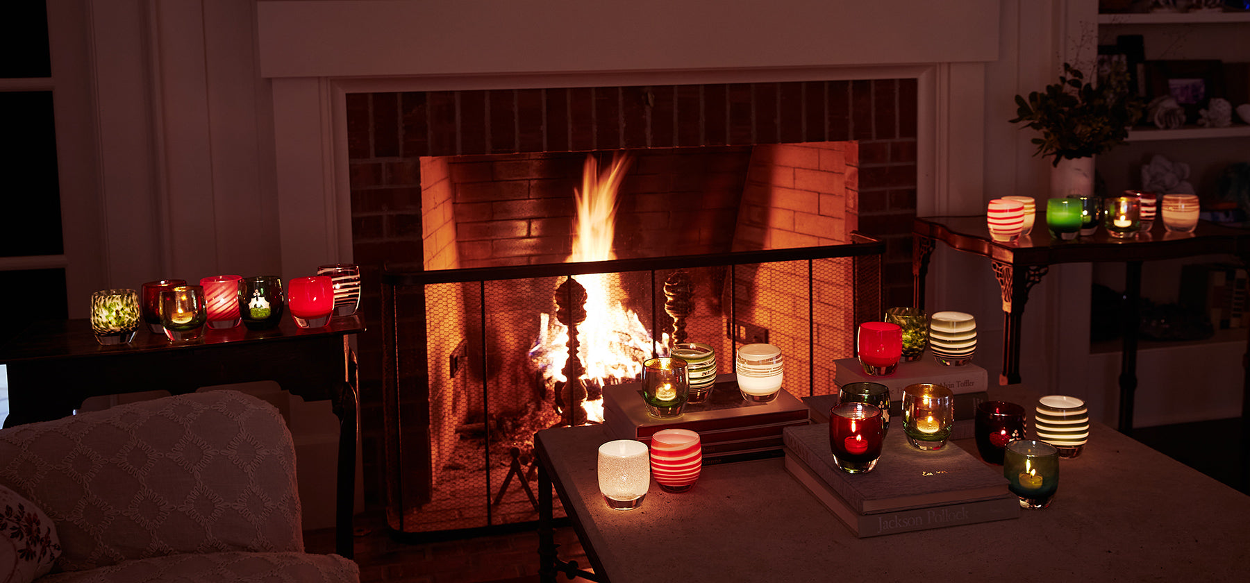 glassybaby holiday collection in a living room: on side tables, coffee table and mantle with lit fire in fireplace.