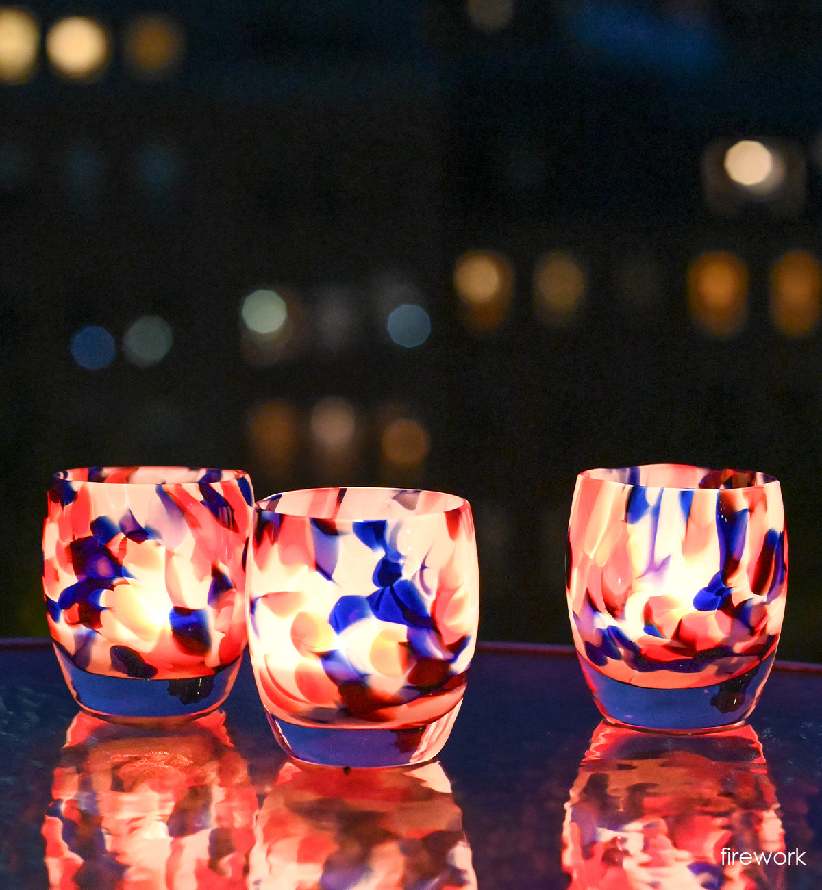 three firework, red white and blue petal, hand-blown glass votive candle holders  on an outdoor reflective surface lit at night.