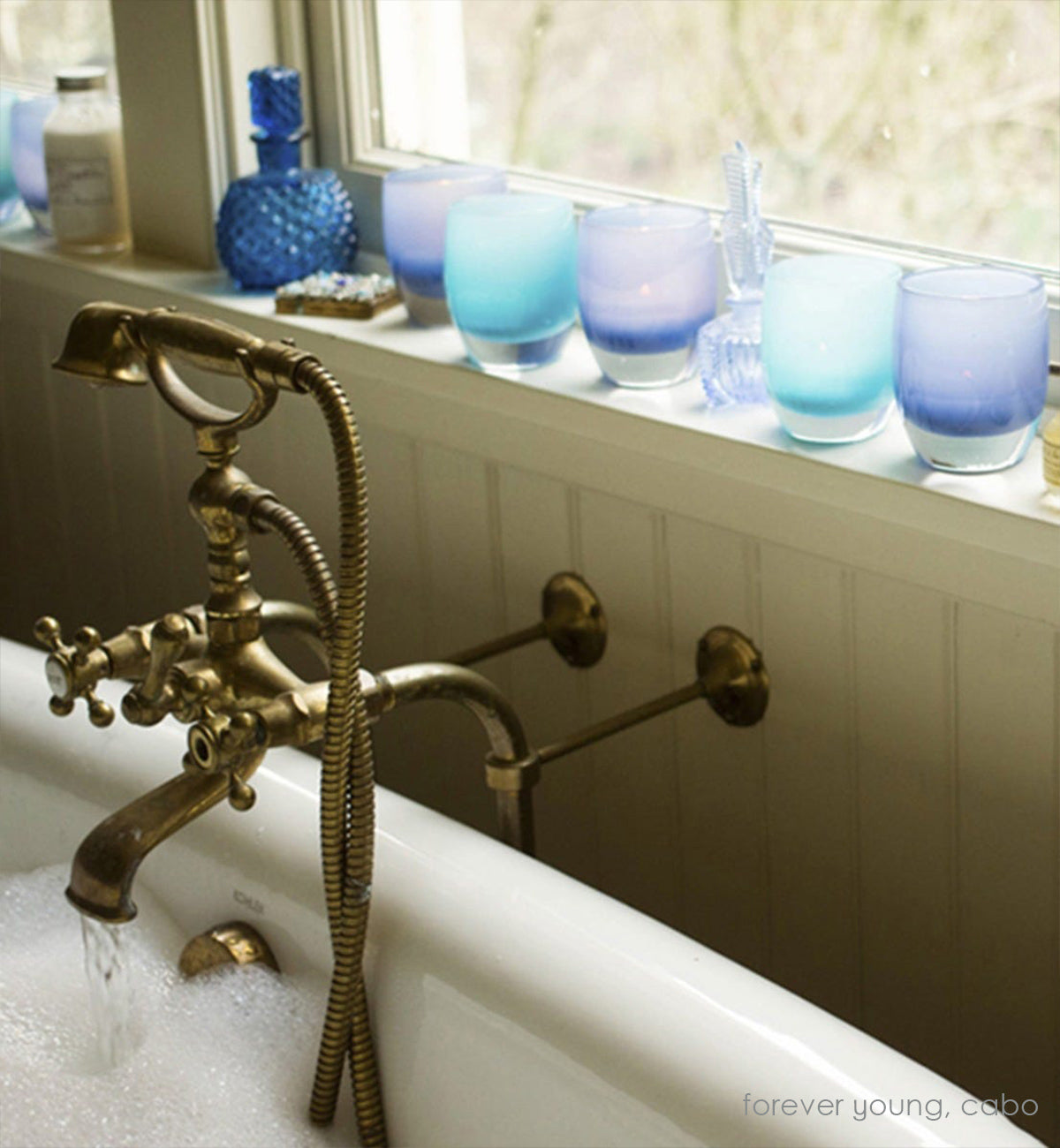 several forever young blue hand-blown glass votive candle holders lined up on a windowsill with cabo, next to a running bubble bath.