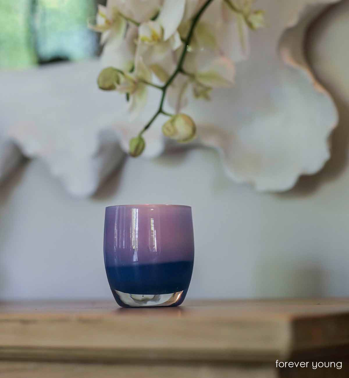 forever young, blue lit with a warm glowing flame on a wooden side table with an orchid bloom and mirror hanging behind. Hand-blown glass votive candle holder.