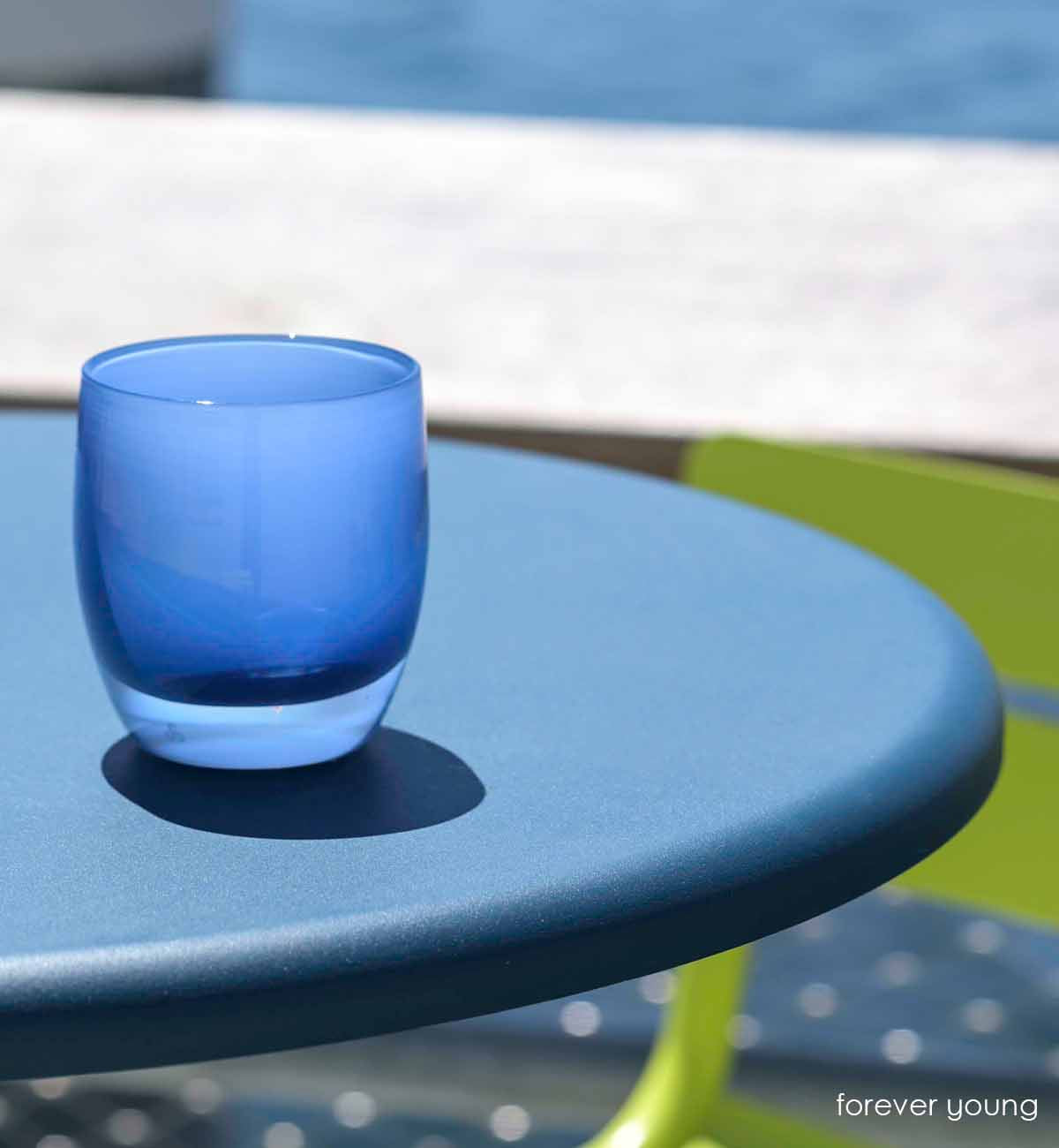 forever young blue hand-blown glass votive candle holder, sunbathing on a table beside a lime green chair, near the water.