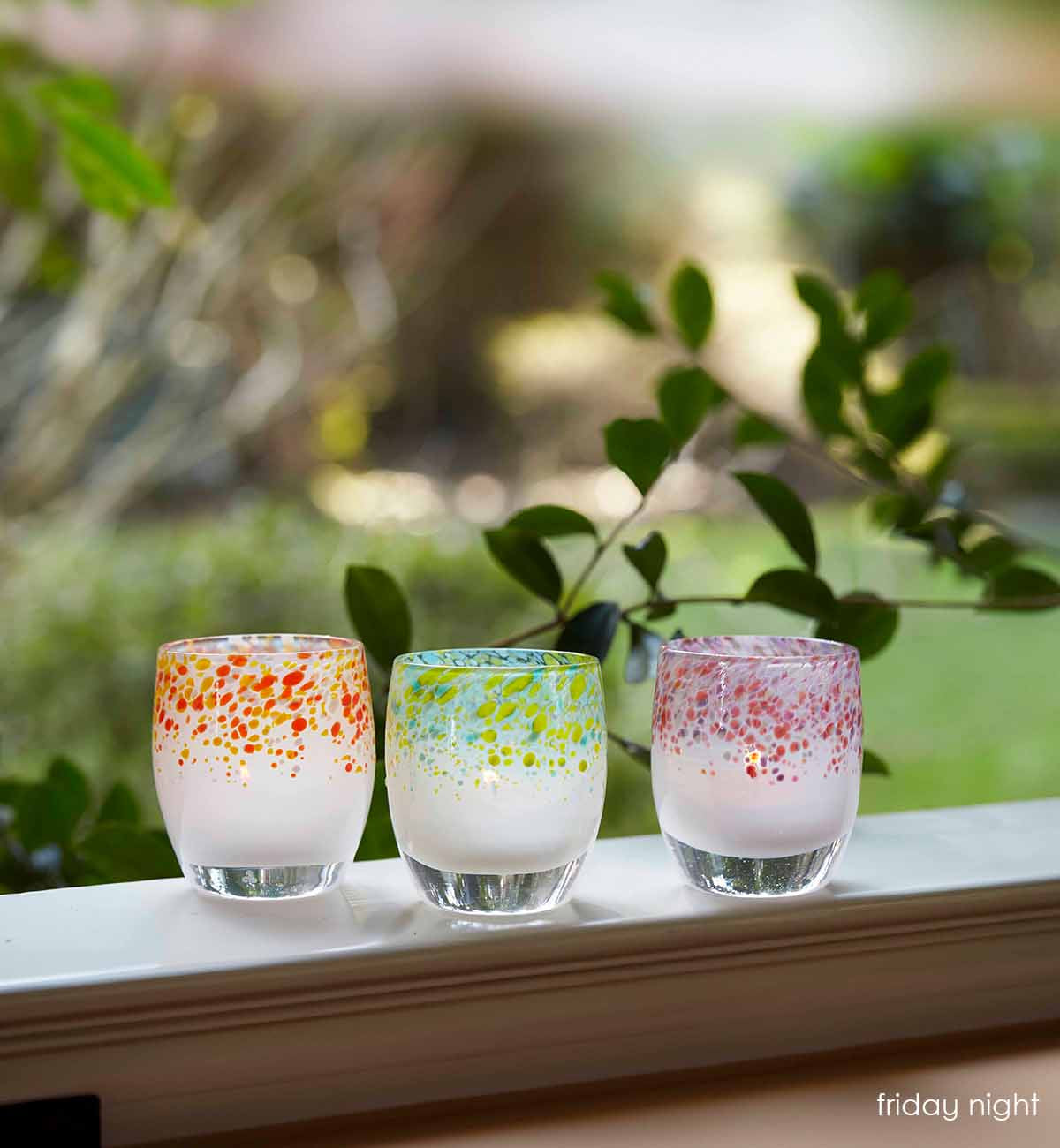 friday night blue green topped hand-blown glass votive candle holder, on a windowsill looking over a green yard.