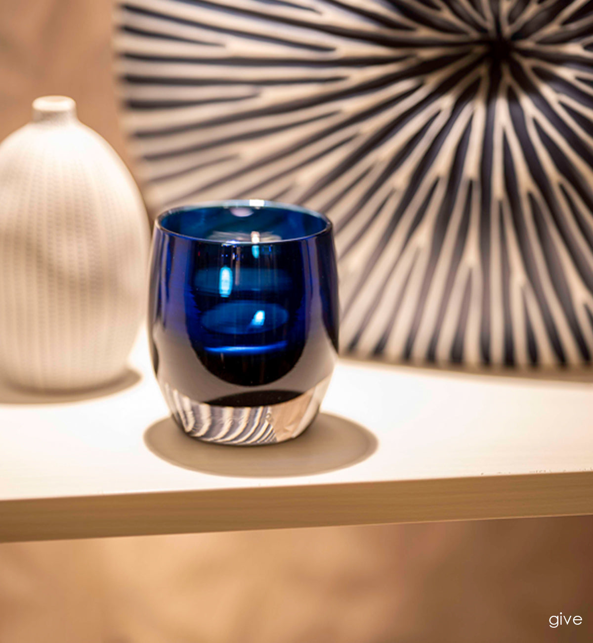 give, hand-blown sapphire blue with silver metallic interior, hand-blown glass votive candle holder lit with a tealight on a creams shelf with a vase and piece of art.