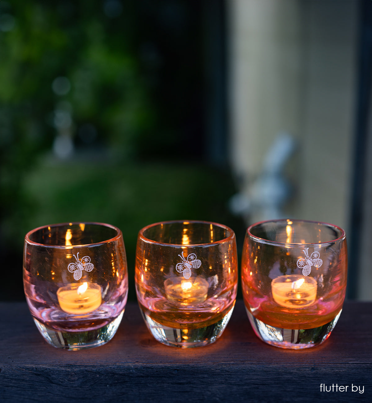 three 'flutter by' transparent rusty orange with engraved butterfly hand-crafted glass candle holders on wood porch railing 