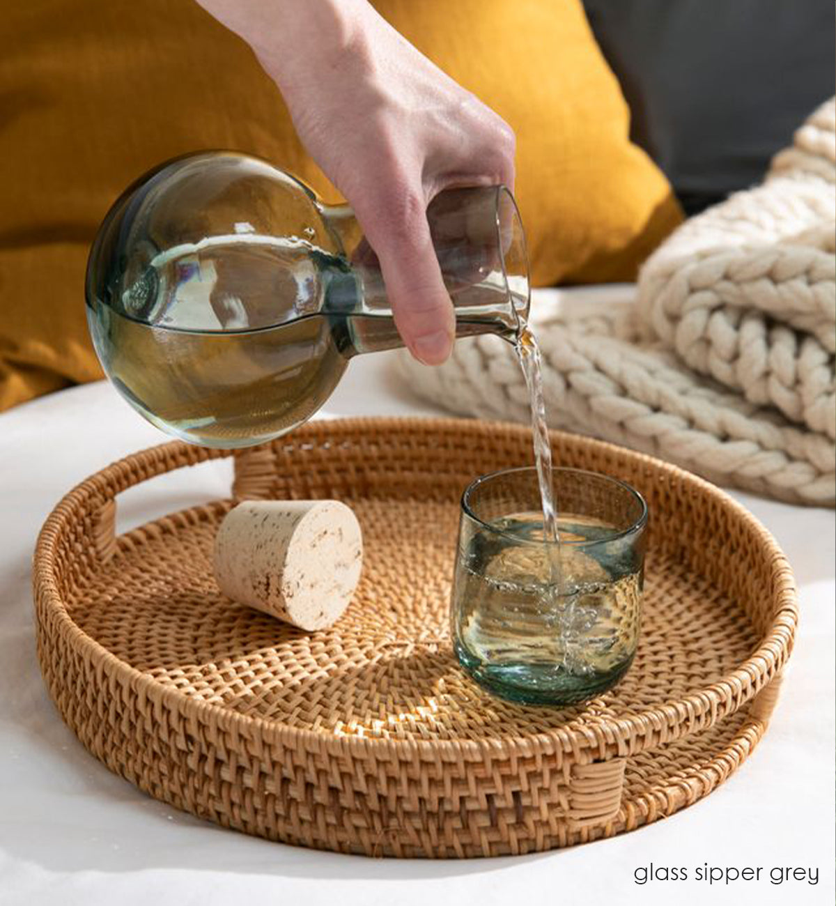 glass sipper grey decanter pouring water into glass sipper grey cup on a woven basket tray with cork on side.