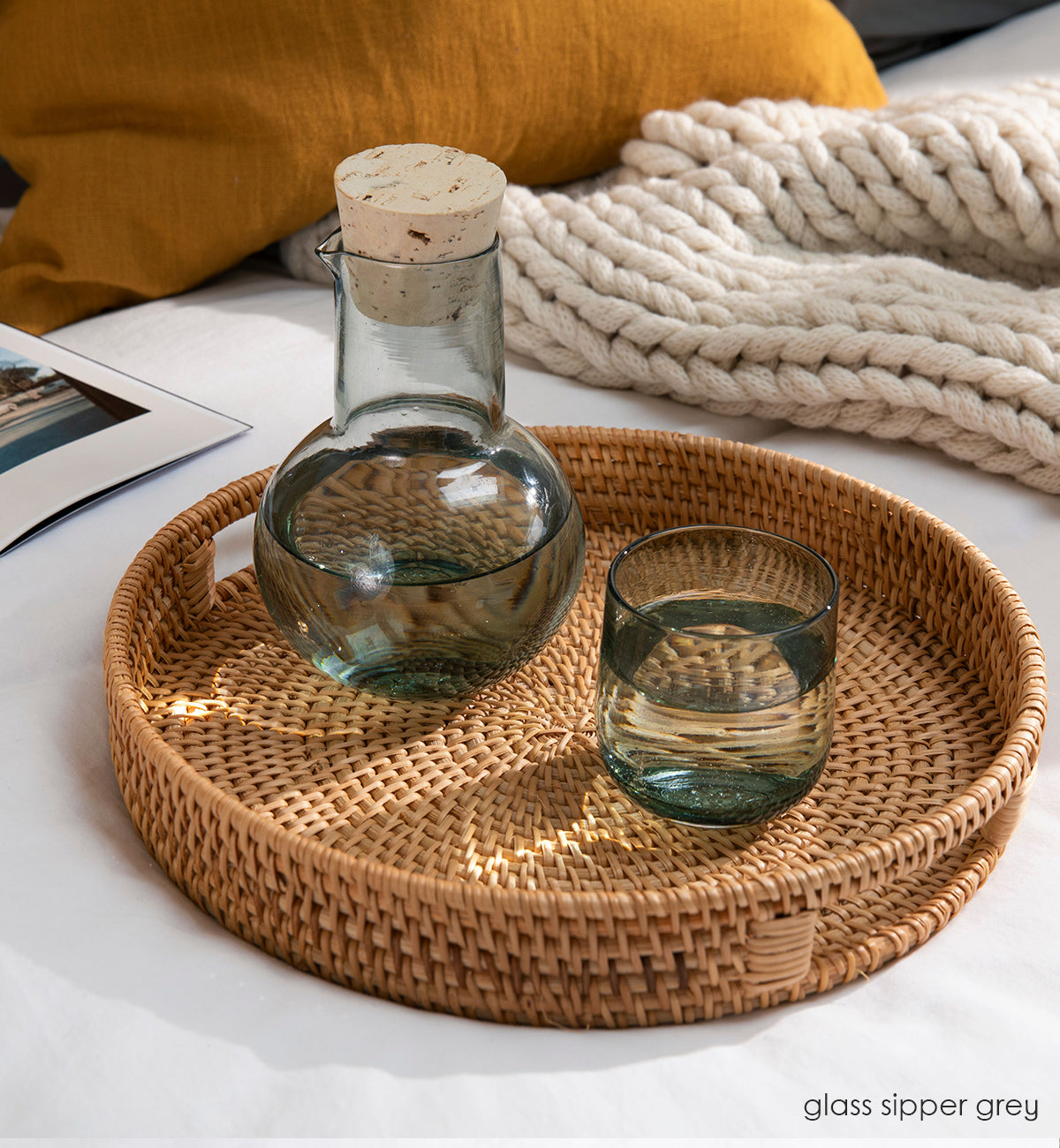 glass sipper grey translucent dark grey hand-blown decanter sitting on a wicker tray filled with water on a cozy couch surrounded by a blanket, pillow, and book.