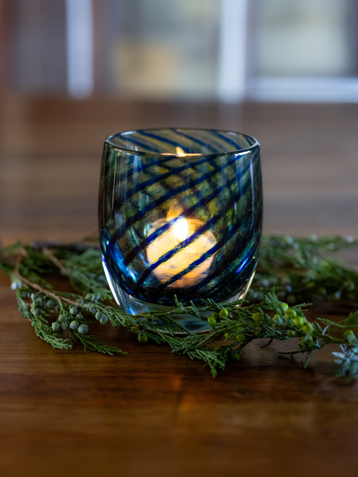 three 'good tidings' blue swirls wrapped around clear glassybaby hand-blown glass candle holder with lit tealight. green garland around it on wood table. 