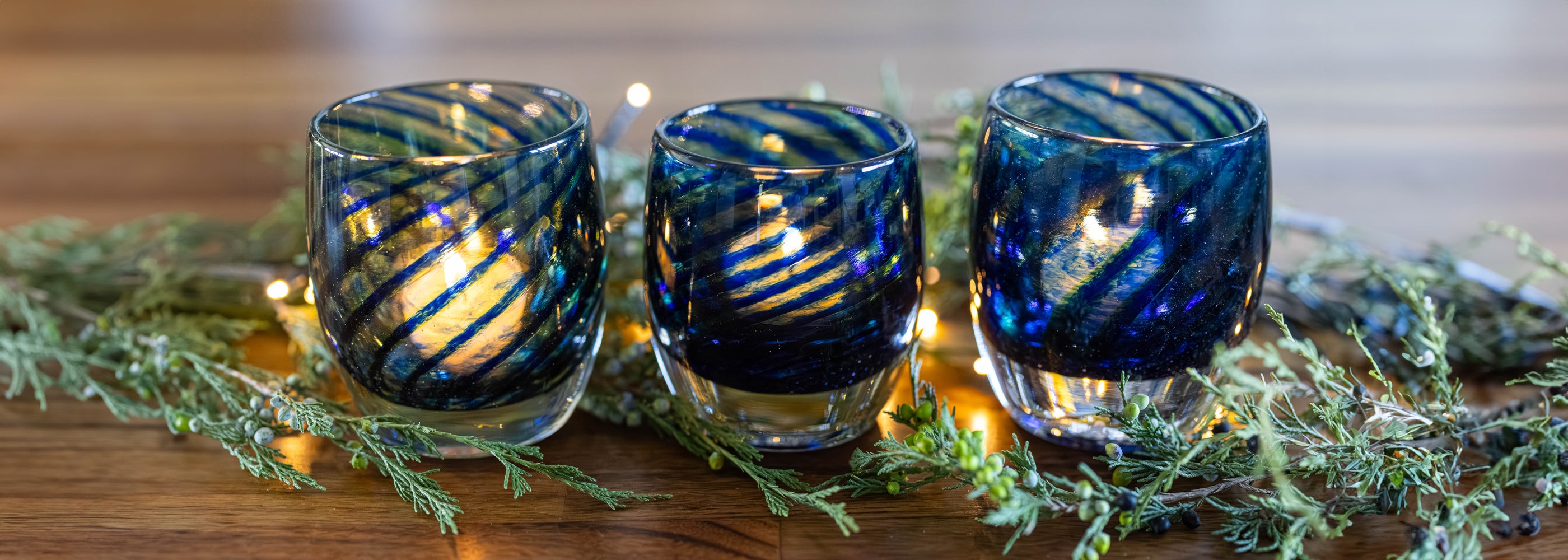 three 'good tidings' blue swirls wrapped around clear glassybaby hand-blown glass candle holder with lit tealight. green garland around it on wood table. 