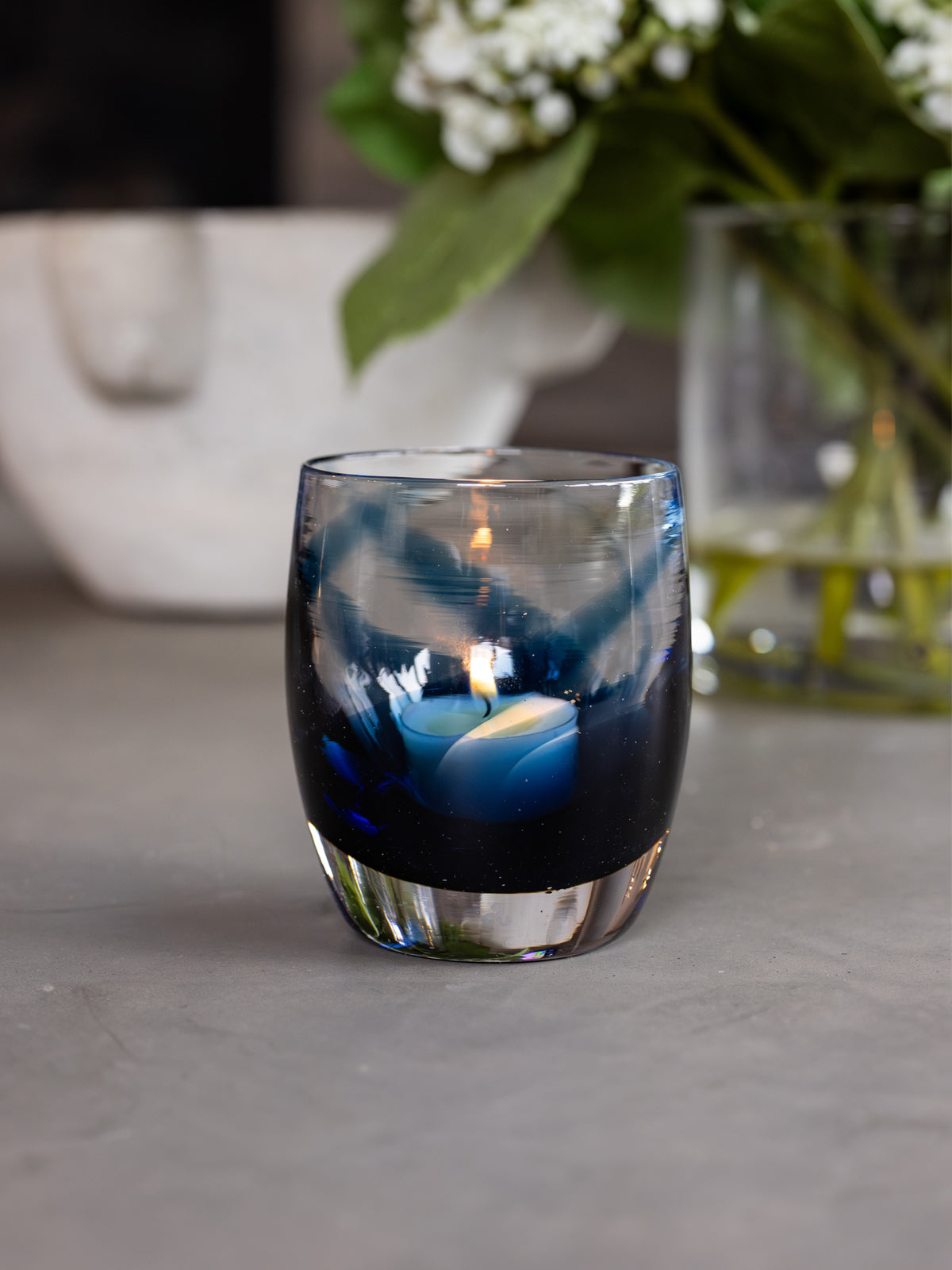 'good vibes' a blue and clear, hand-blown glass candle holder on gray granite table with a white bowl and clear vase with white flowers in the back ground 