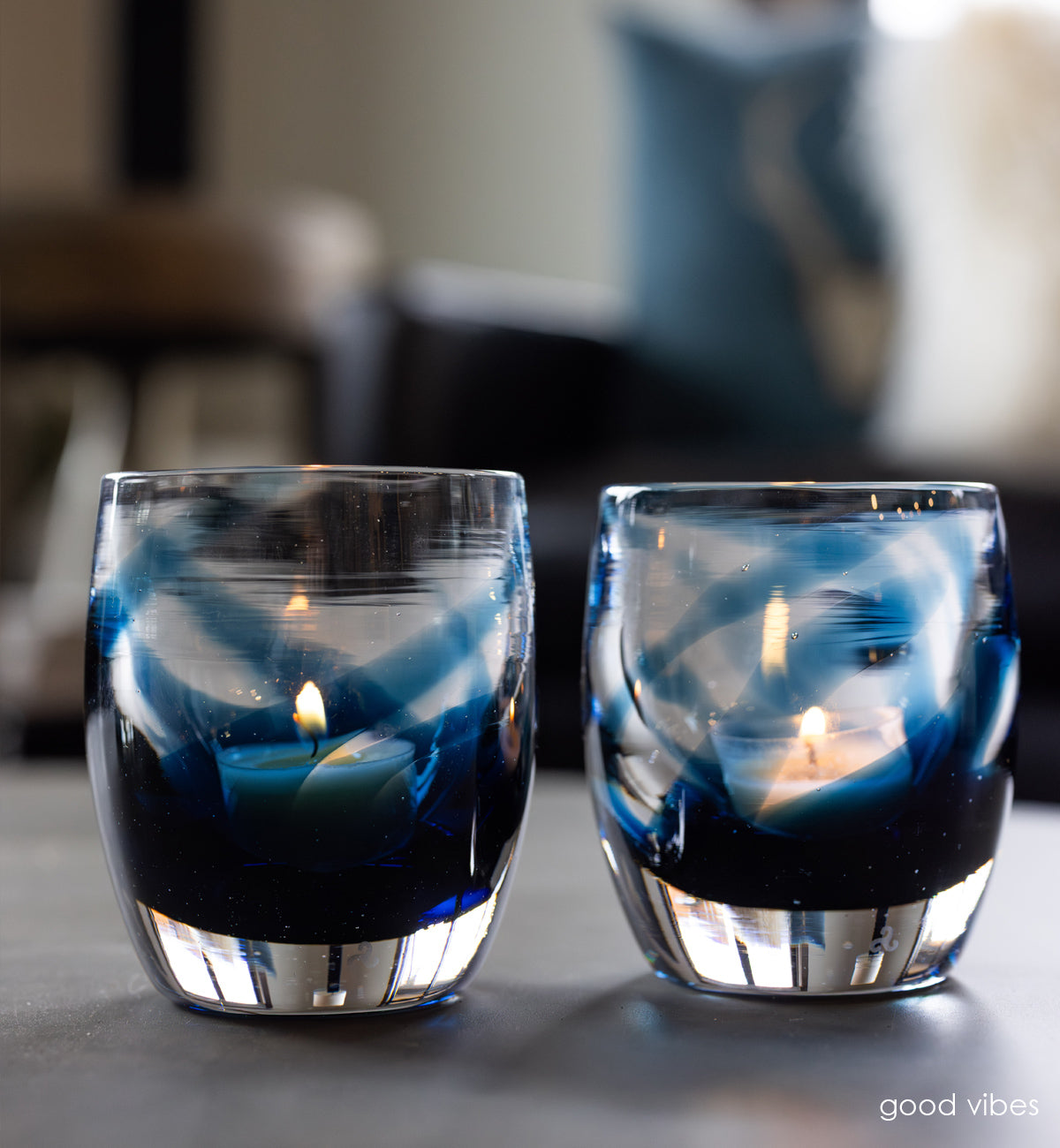 'good vibes' a blue and clear, hand-blown glass candle holder on gray granite table with a couch in the background with blue and white pillows