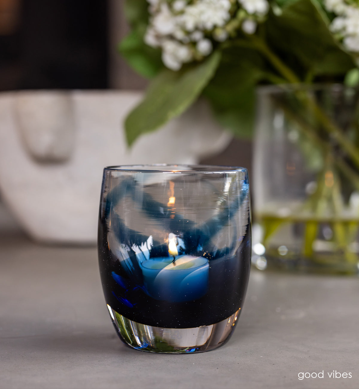 'good vibes' a blue and clear, hand-blown glass candle holder on gray granite table with a white bowl and clear vase with white flowers in the back ground 