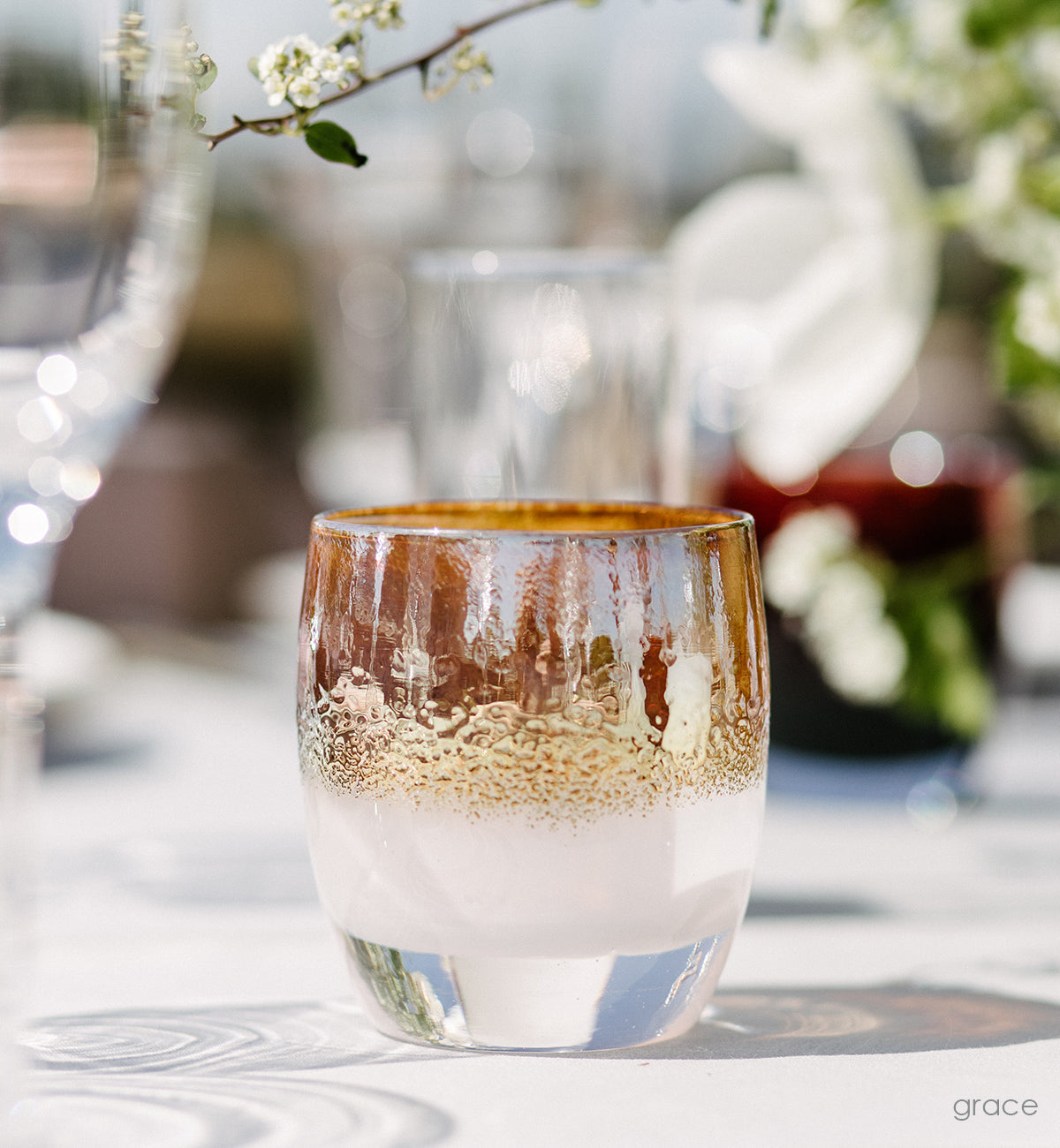 grace golden topped white hand-blown glass votive candle holder on a white table in the sun with drinking glasses and flowers in background.
