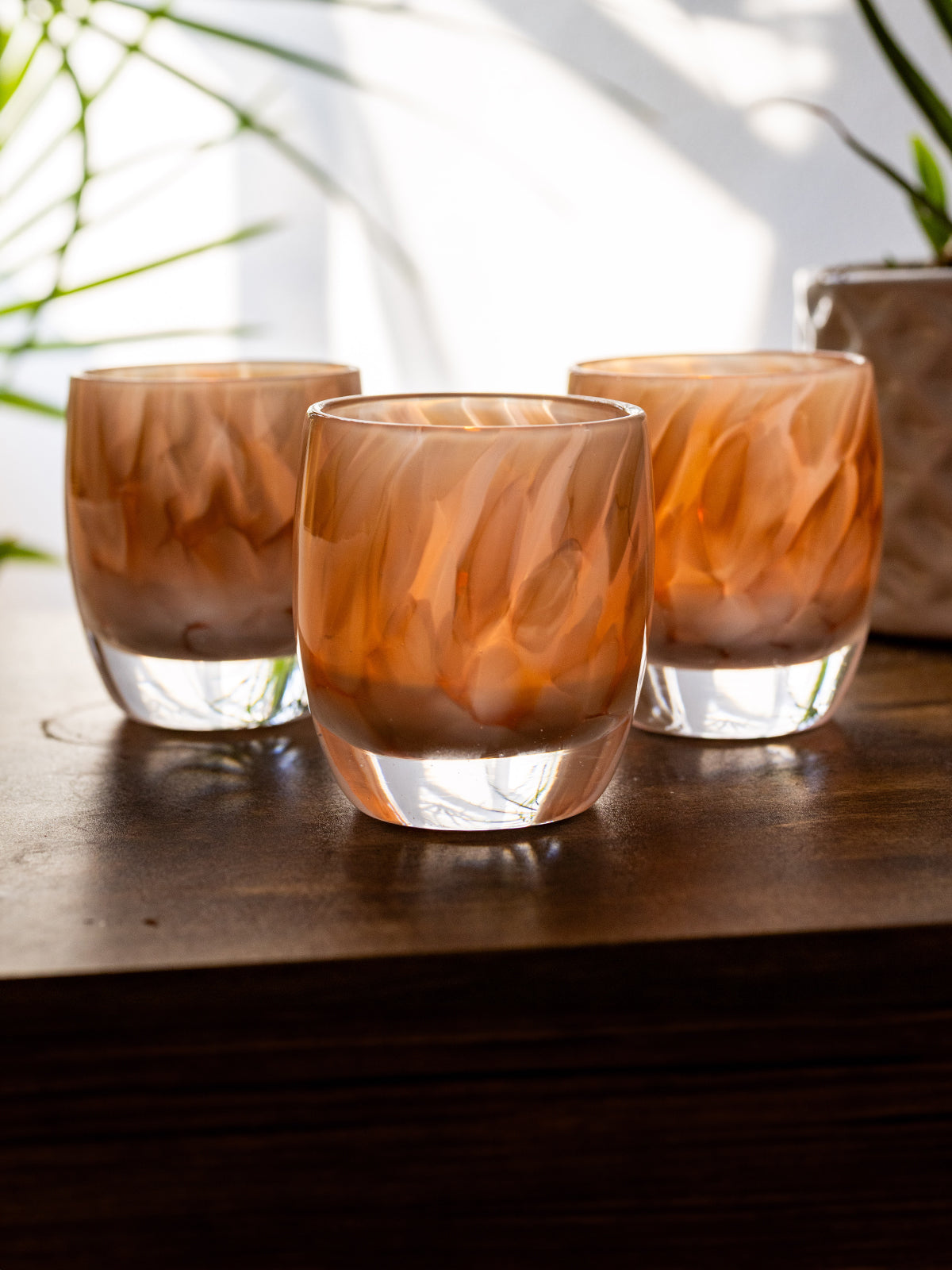 group of 'gracious' white and brown petal, hand-blown glass votive candle holder when lit gives off an orange glow on wood desk. green plants in background against white wall. 