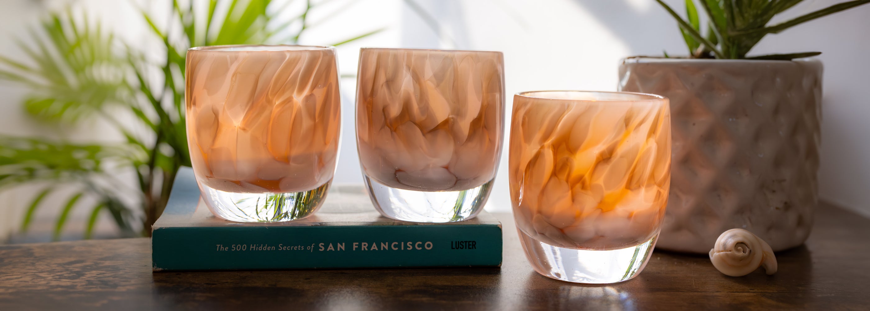 group of 'gracious' white and brown petal, hand-blown glass votive candle holders when lit gives off an orange glow on wood desk with a book. green plants in background against white wall. 