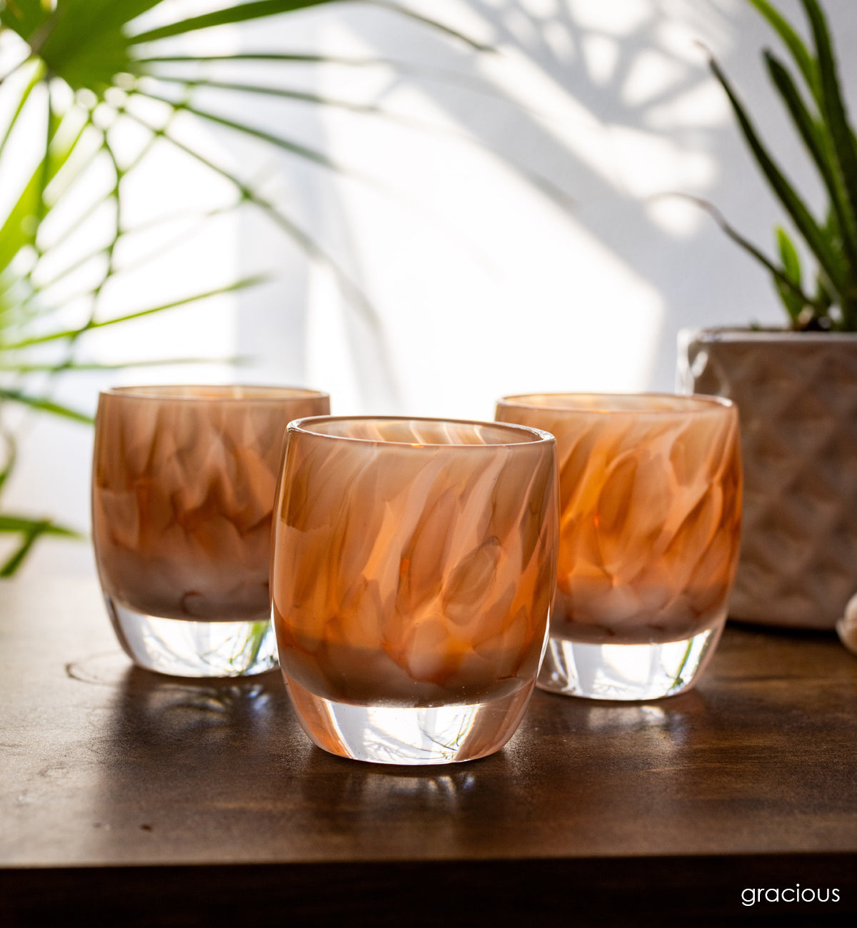 group of 'gracious' white and brown petal, hand-blown glass votive candle holder when lit gives off an orange glow on wood desk. green plants in background against white wall. 
