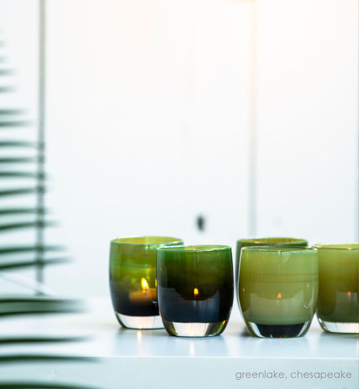 greenlake, olive green hand-blown glass votive candle holders with chesapeake on a white table in a light setting with a green plant off to the side.