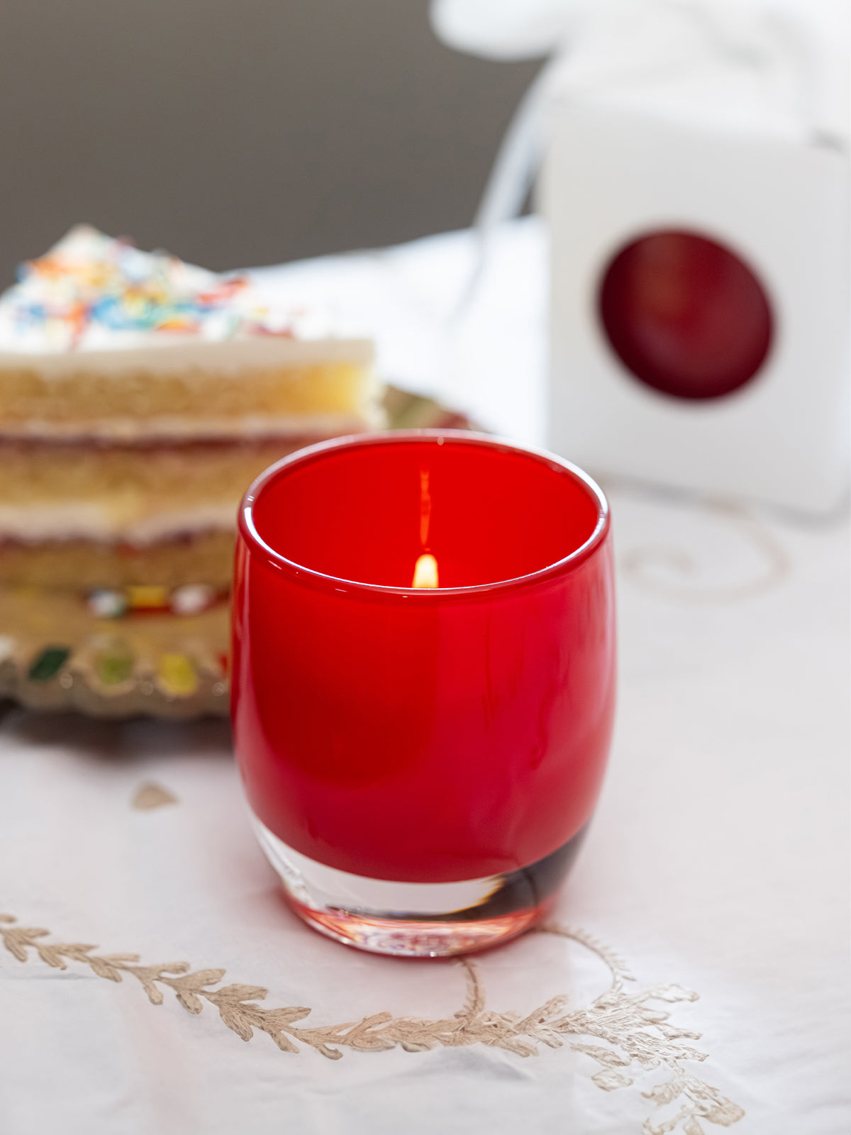 Red hand-blown glass candle holder with a slice of cake on a patterned tablecloth