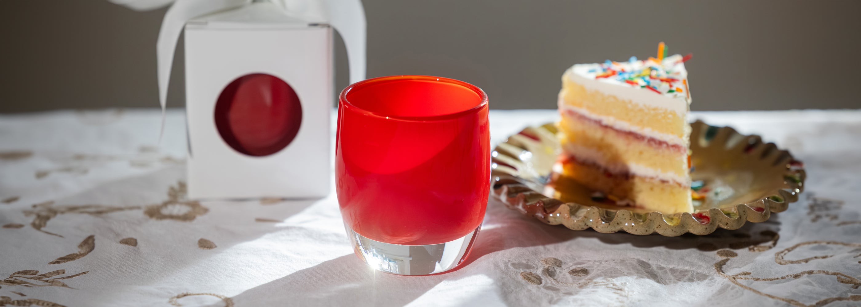 Red hand-blown glass candle holder with a slice of cake on a patterned tablecloth