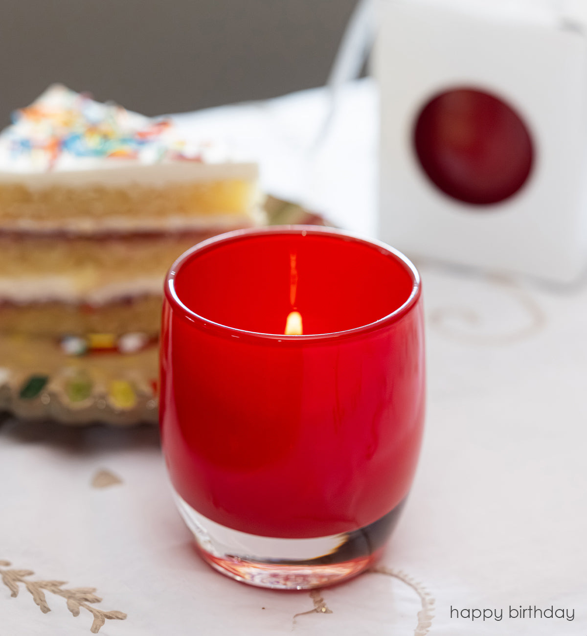 happy birthday bright red, hand-blown glass votive candle holder with a blurred birthday cake in the background