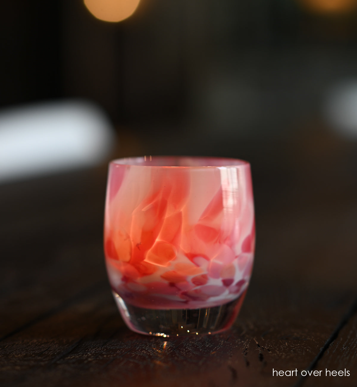 heart over heels white and pink speckled hand-blown glass votive candle holder on a dark wood table.