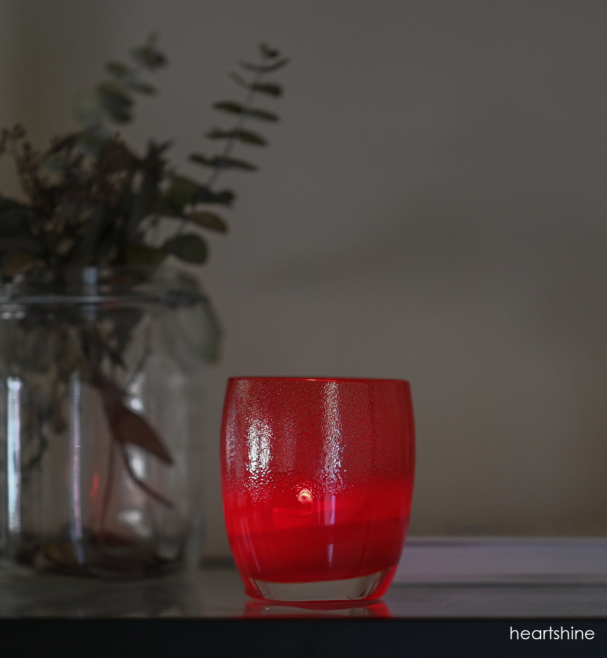 heartshine, shimmering glitter that enrobes the top of a bright red base, hand-blown glass votive candle holder on a dark table with cream wall behind and a vase of green leafed stems.