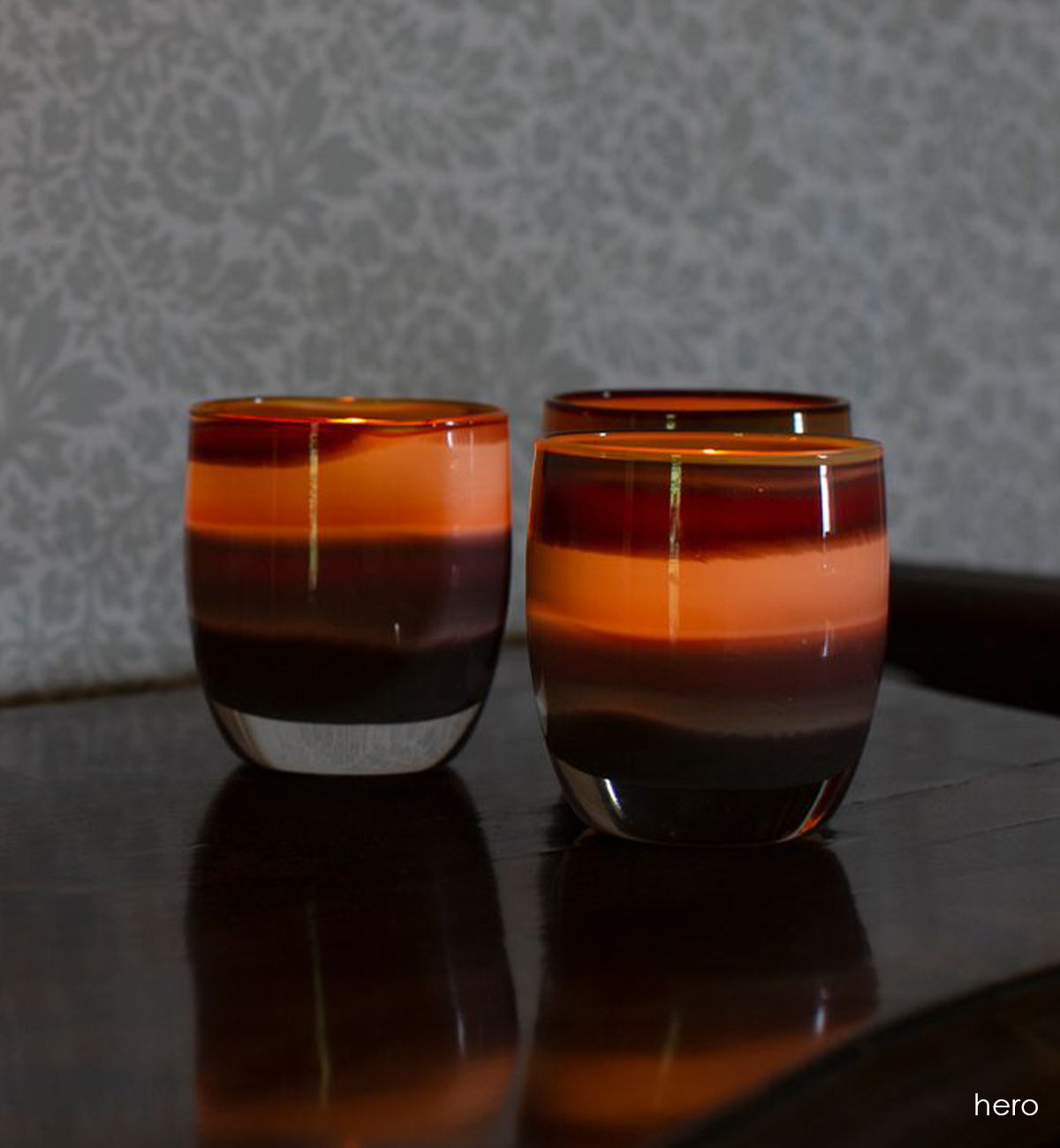 three hero multi brown hand-blown glass votive candle holders on a wood surface in front of gray and white wallpaper.