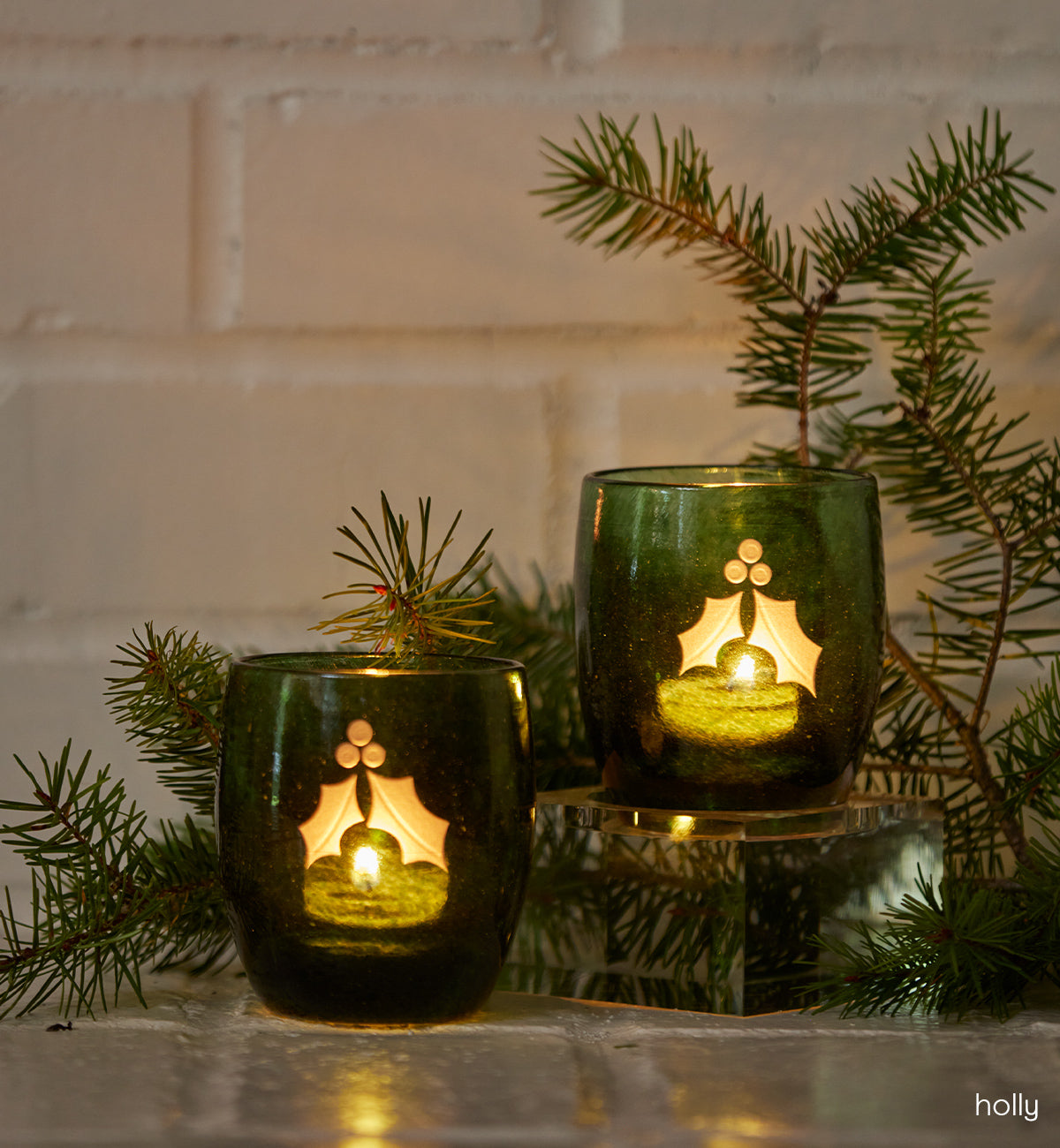 two holly, dark green with etched holly leaf and berries, hand-made glass votive candle holder sitting on a table with some green sprigs.