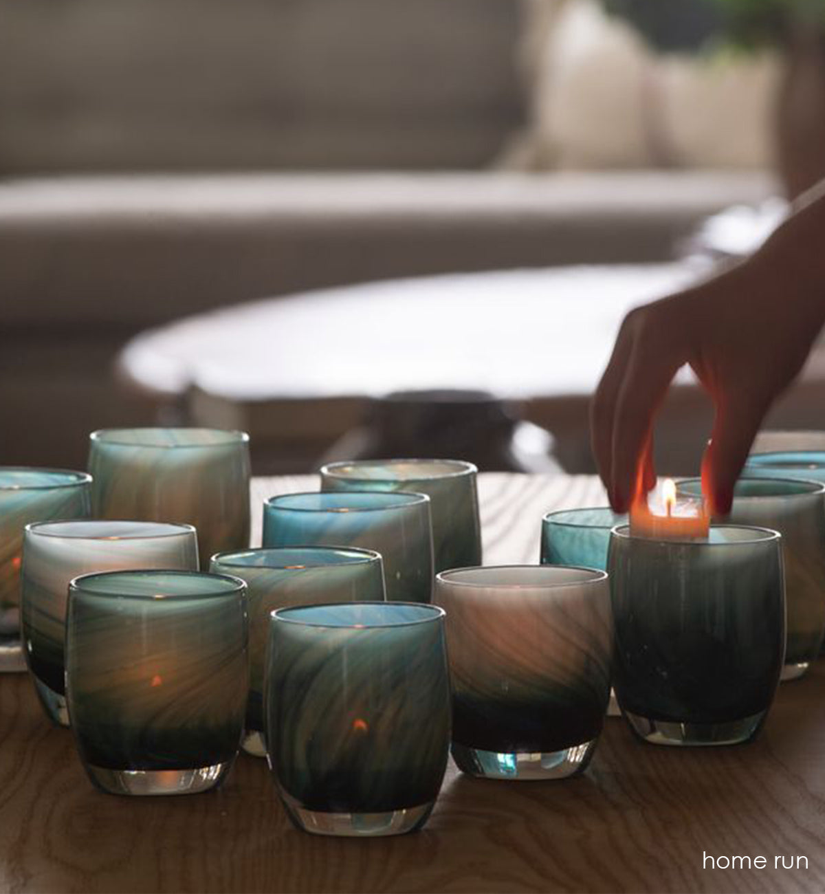 grouping of home run, multi-colored blue swirl hand-blown glass votive candle holders on a light wood coffee table, with a tealight being placed inside one.