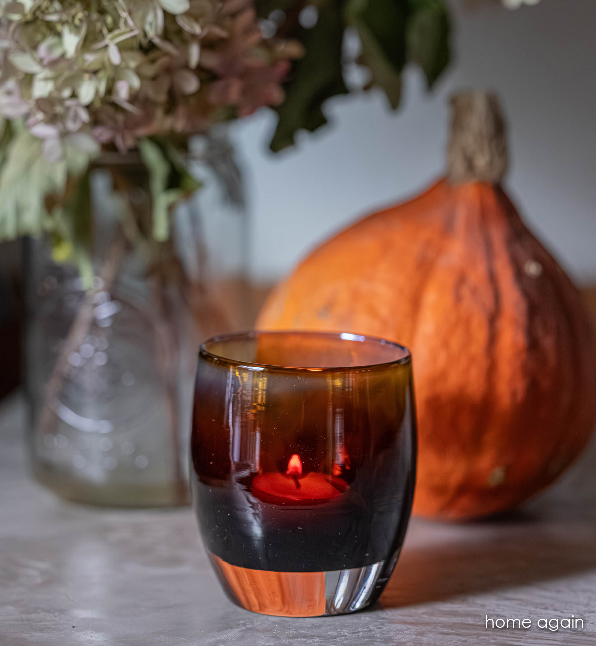 home again glassybaby: brown gradient hand-blown glass votive candle holder with a gourd and clear flower vase behind it. 