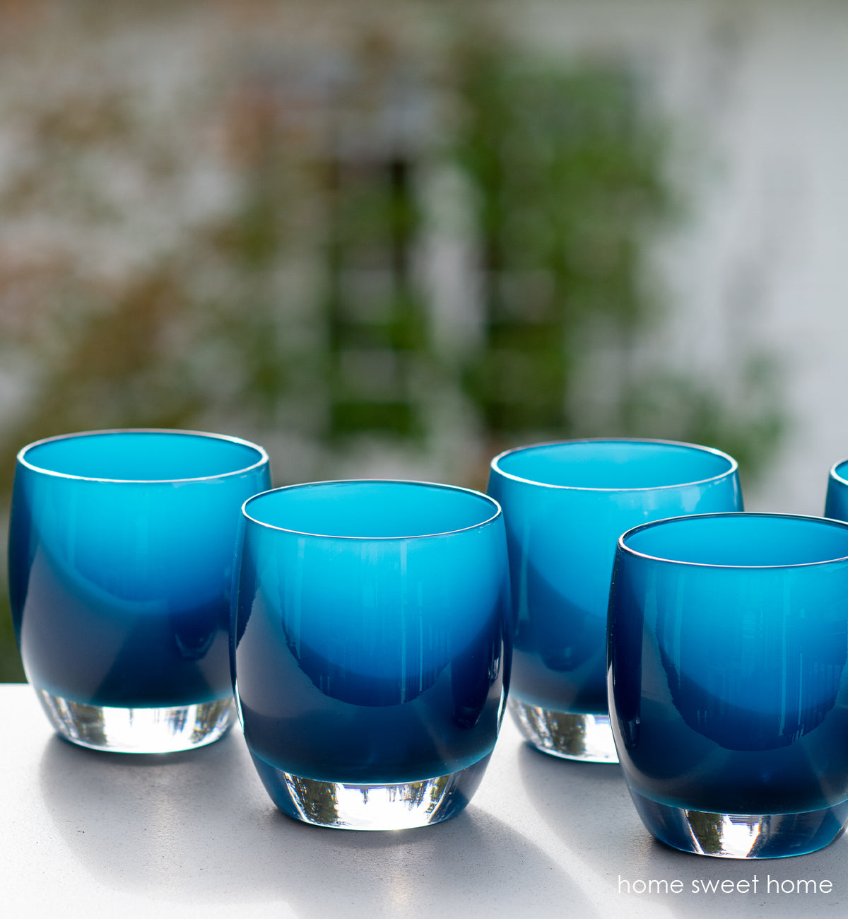 several home sweet home deep blue, hand-blown glass votive candle holders side by side on a surface overlooking a backyard.