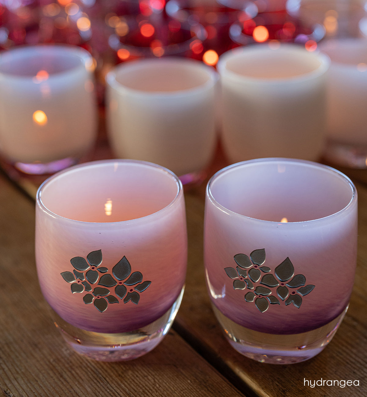 Two pink glass candle holders with with hand etched hydrangea design on a wooden surface.