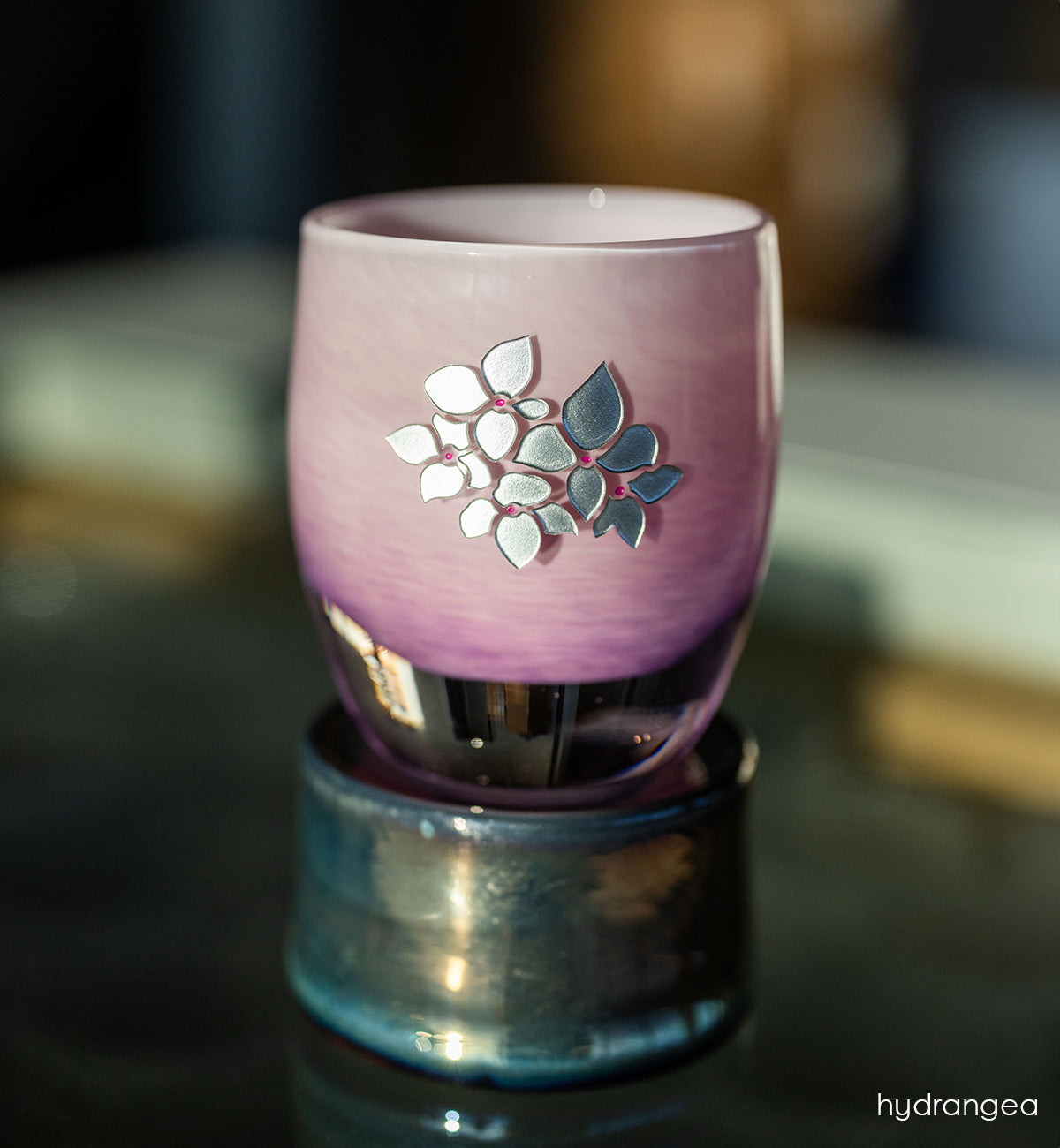 Pink glass candle holder with hand etched hydrangea design on a reflective surface.