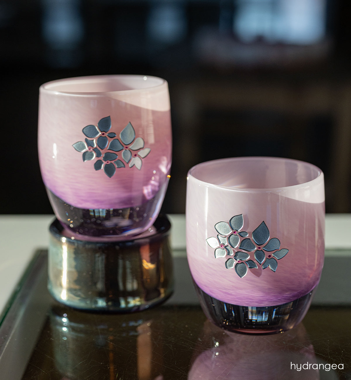 Two pink glass candle holders with with hand etched hydrangea design on a reflective surface.