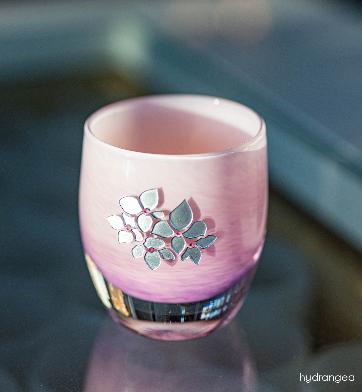 Pink glass candle holder with hand etched hydrangea design on a reflective surface.