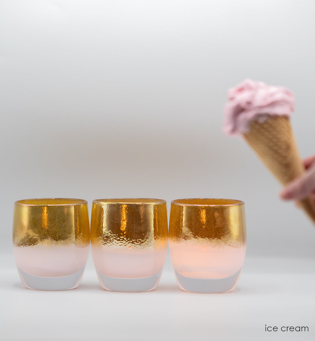 three ice cream pink gold hand-blown glass votive candle holders placed against a white background with a blurred ice cream cone in corner.