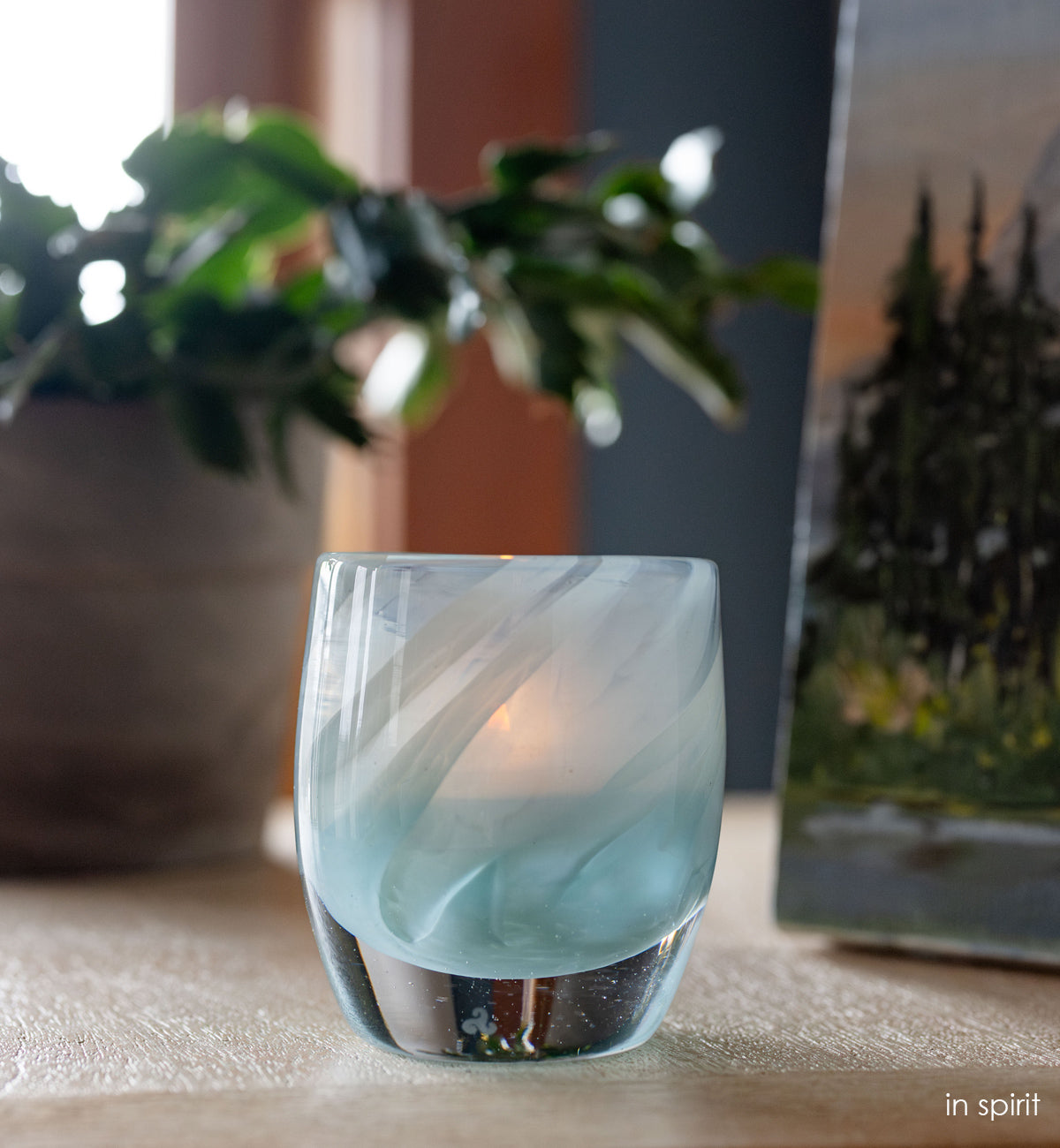 'in spirt' light blue hand-blown glass candle holder with green plant and painting in background.