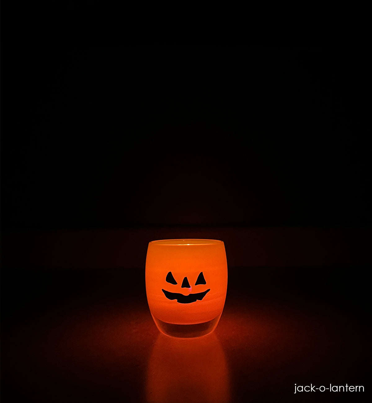 jack-o-lantern, orange hand-blown glass votive candle holder with a spooky jack-o-lantern face hand-etched in black fill sitting and glowing in a dark setting.