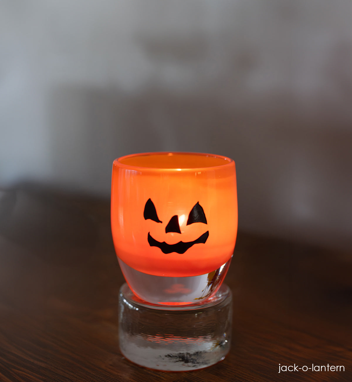 jack-o-lantern, orange hand-blown glass votive candle holder with a spooky jack-o-lantern face hand-etched in black fill on clear pedestal. 
