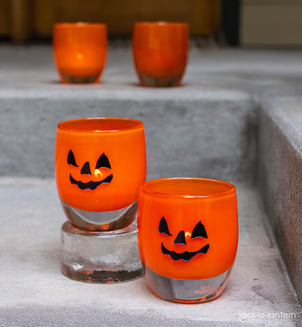 two jack-o-lantern, orange hand-blown glass votive candle holders with a spooky jack-o-lantern face hand-etched in black fill one on clear pedestal. 
